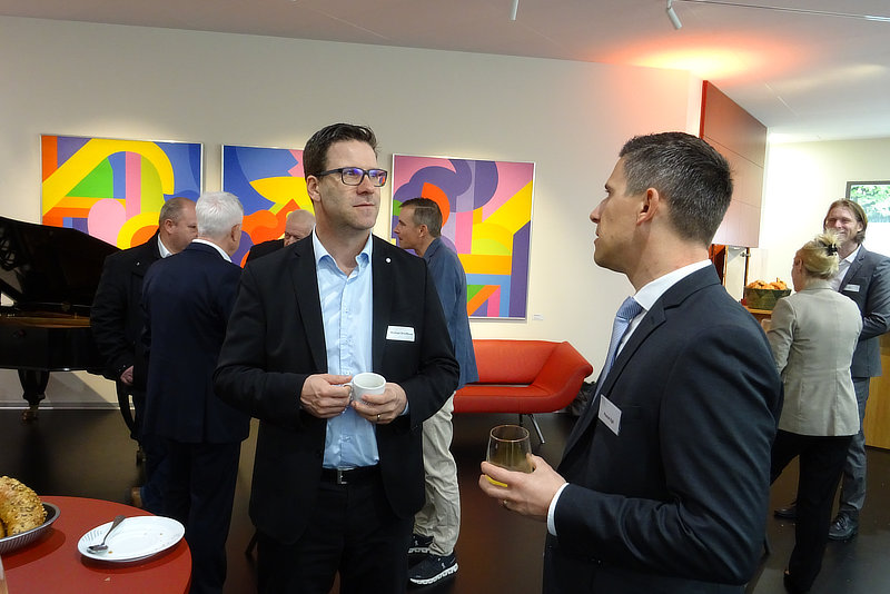 Zwei Männer im Anzug unterhalten sich in einer modernen Umgebung mit bunten Wandbildern, während im Hintergrund weitere Personen und ein Klavier zu sehen sind.